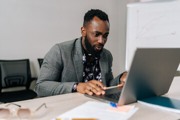Successful black male businessman sitting at worktable at modern office, typing on computer...