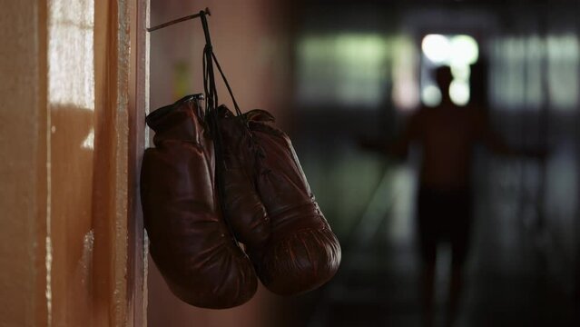 The shot shows a close up of a doorjamb with a rusty nail nailed to it on which brown boxing gloves hang. In the background is a man jumping rope, he is doing cardio, working out. Before the fight