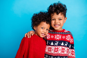 Portrait of two funny little friends with wavy hair dressed red ornament sweaters embrace on new year isolated on blue color background - obrazy, fototapety, plakaty