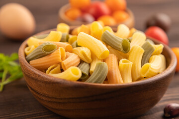 Rigatoni colored raw pasta with tomato, eggs, spices, herbs on brown wooden. Side view, selective focus