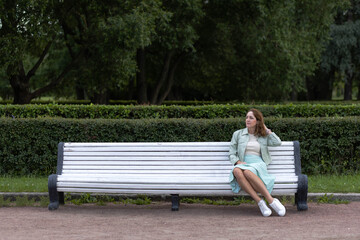 The woman sits alone on a long white bench in the park. Loneliness.