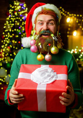 Merry Christmas and Happy New year. Bearded man in Santa hat with decorated beard holds Christmas gift. Santa delivery man with stylish decorated beard with New year present. Christmas beard style.