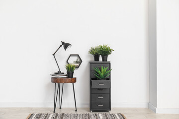 Desk lamp on small table and file cabinet with houseplants near white wall - obrazy, fototapety, plakaty