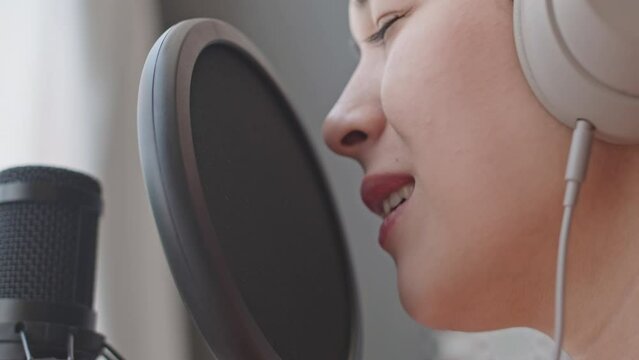 Close up shot of young female singer in headphones singing in microphone while recording song at home studio