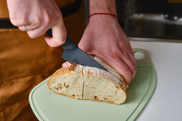 Man cuts a piece of fresh bread with a knife and makes a delicious cheese sandwich in the kitchen
