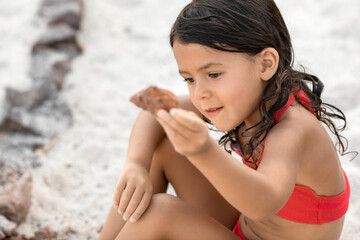 Little toddler girl in red swimsuit sitting on sandy beach and playing with rock at summer vacation