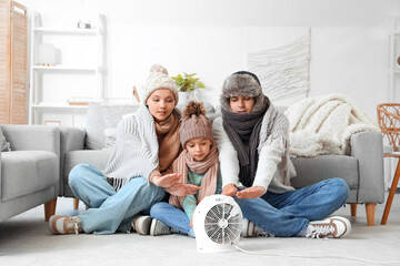 Frozen family in winter clothes warming near electric fan heater at home - obrazy, fototapety, plakaty
