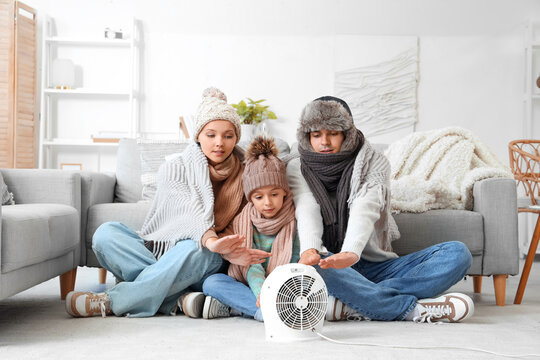 Frozen family in winter clothes warming near electric fan heater at home