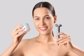 Beautiful young woman with facial massage tool and cream on light background