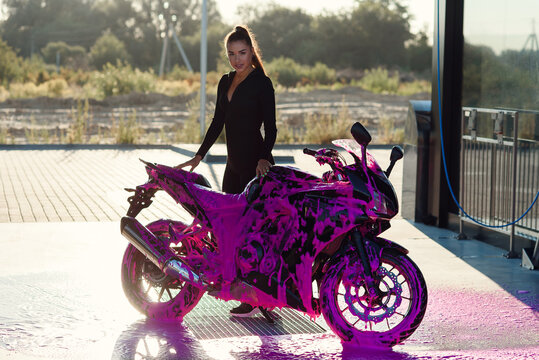 Pretty girl in black seductive suit stands near motorcycle at self-service car wash at sunrise.