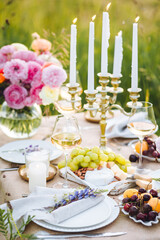 Romantic sunset wedding table decor for a couple with vintage chandelier, burning candles, snacks, white wine, fruits, ranunculi flowers. Summer vibes, field or meadow, outdoors event