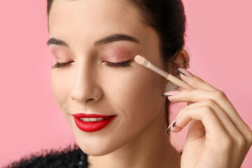 Beautiful young woman with brush applying eyeshadows on pink background