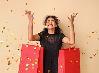 Beautiful African-American woman with shopping bags and confetti on beige background. Black Friday...