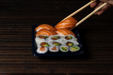 Sushi To Go. Holding Chopsticks with Salmon Nigiri. Dark Mood Sushi Tray on Wooden Background.