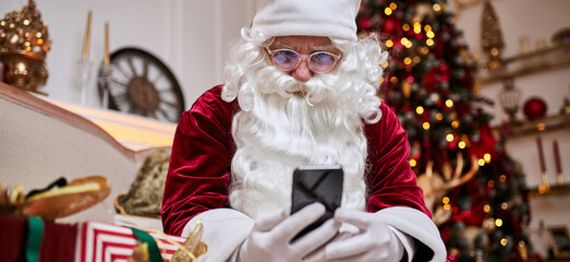 Santa Claus use mobile phone sitting neat christmas tree with gifts. New year and Merry Christmas  - 670742381
