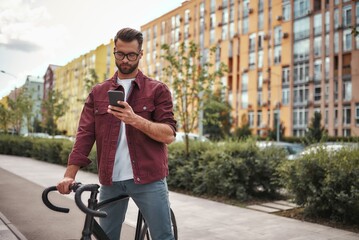 Chatting with friend. Handsome man with stubble in casual clothes and eyeglasses holding mobile phone while walking with his bicycle outdoors