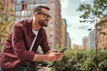 Good news. Side view of handsome man with stubble in casual clothes and eyeglasses leaning at his bicycle, holding mobile phone and smiling while standing outdoors - obrazy, fototapety, plakaty