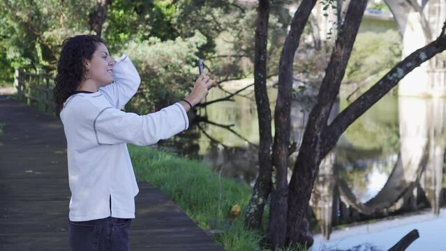 Young girl taking a selfie while grooming herself in a natural environment with water in the background. Brown curly hair with white sweatshirt. Concept of social networks and young people.