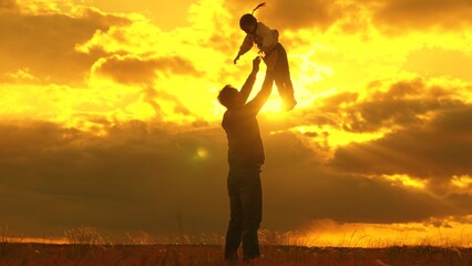 Happy dad plays with his baby daughter, throws child into sky with his hands, happy child laughs. Silhouette, father, child playing together in park against backdrop of sun clouds. Family, dream fly