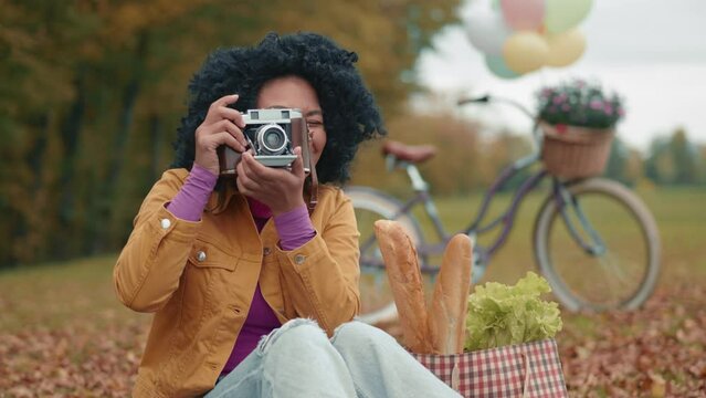Attractive smiling colored woman taking pictures on vintage camera sitting on yellow blanket of fallen leaves in autumn park after shopping, cycling. High quality 4k footage