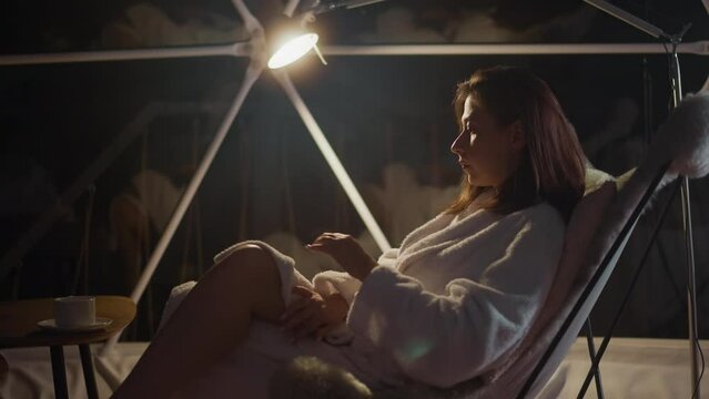 Gloomy lady covers legs with terry robe sitting in armchair by light of lamp near panoramic window at night. Lack of support leading to suffering from loneliness slow motion