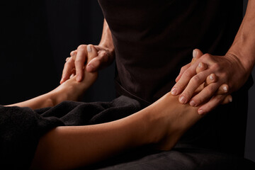 handsome male masseur doing a massage on a girl's leg on a black background, concept of therapeutic relaxing massage - obrazy, fototapety, plakaty