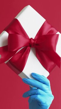 Vertical video. Valentine Day gift. Pandemic holidays. Hand in blue protective glove holding wrapped present in white box with ribbon bow isolated on red background.