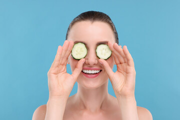 Beautiful woman covering eyes with pieces of cucumber on light blue background