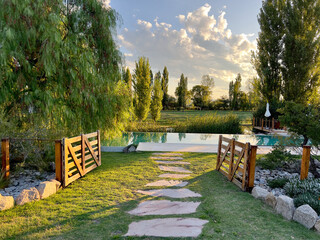 Peaceful garden scene with an open wooden bridge to a pool and a stone path. Sun setting behind the...