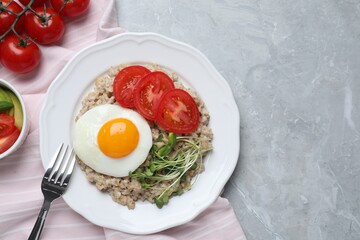 Delicious boiled oatmeal with fried egg, tomatoes and microgreens on light grey marble table, flat lay. Space for text