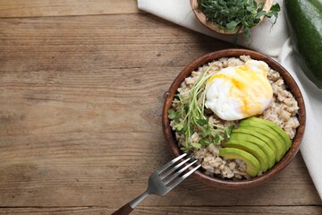 Delicious boiled oatmeal with poached egg, avocado and fork on wooden table, flat lay. Space for text