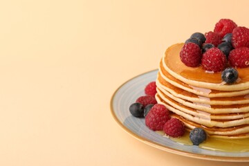 Stack of tasty pancakes with raspberries, blueberries and honey on pale orange background, closeup. Space for text