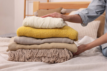 Woman with stack of different folded blankets and clothes on bed in room, closeup. Home textile