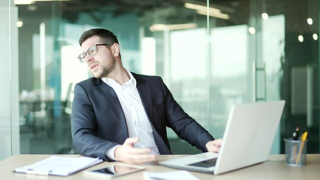 Disappointed stressed businessman in a shirt reads bad news on a laptop while sitting at workplace in business office. Frustrated entrepreneur is angry reviewing negative information on the computer