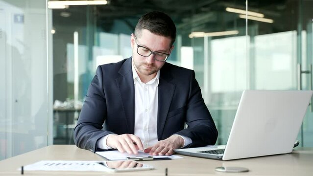 Confident businessman is signing documents while sitting at a desk at a workplace in a business office. Serious boss owner in a formal suit rereads the folder with documentation and signs the contract