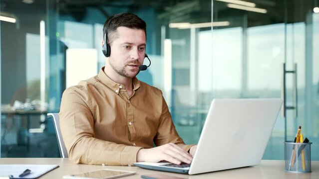 A corporate operator who works in customer support speaks on a video call using a laptop computer. Call center agent in wireless headset helping clients with complaints in office. Online consultation
