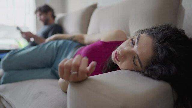 Exhausted young woman laid on couch with tired expression while boyfriend is in background looking at phone. Lonely people alone together