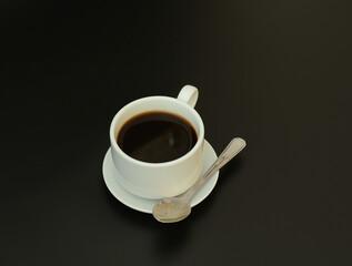 White porcelain cup of hot black coffee on a saucer with a spoon on a black abstract background.