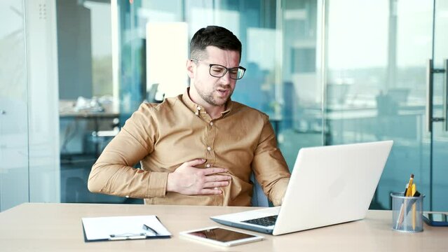 Young businessman feels stomach pain while working on a laptop sitting at workplace in business office. Sick worker has heartburn, gastritis or poisoning. Man suffers from spasms and is constipated