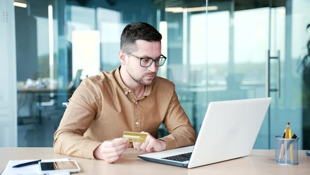 Shocked businessman checks his credit card on laptop while sitting at workplace in business office. Frustrated sad male faced fraud, money was stolen from his account. He became a victim of deception