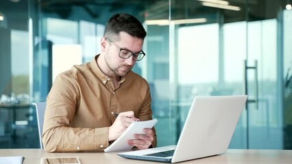 Businessman watching video call training notes in notebook looking at laptop computer screen while sitting at desk at workplace in business office. Worker on remote certification training. Skill up