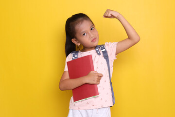 Happy Asian little girl holding book and carrying backpack showing strong hand gesture over yellow...
