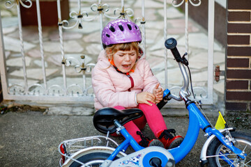 Obraz premium Cute little girl sitting on the ground after falling off her bike. Upset crying preschool child with safe helmet getting hurt while riding a bicycle. Active family leisure with kids.