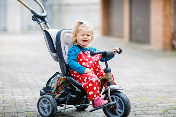 Cute adorable toddler girl sitting on pushing bicycle or tricycle. Little baby child going for a walk with parents cold cloudy day. Happy healthy kid in colorful clothes - obrazy, fototapety, plakaty