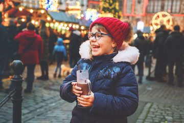 Little cute preschool girl drinking hot children punch or chocolate on German Christmas market. Happy child on traditional family market in Germany, Laughing kid in colorful winter clothes - obrazy, fototapety, plakaty