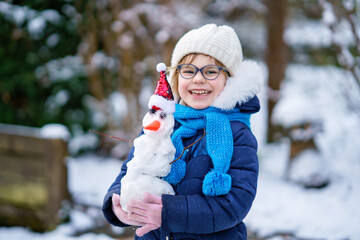 Cute little preschool girl with glasses making mini snowman. Adorable healthy happy child playing and having fun with snow, outdoors on cold day. Active leisure with children in winter
