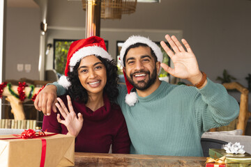Happy biracial couple with santa hats embracing and waving hands among gifts at christmas at home