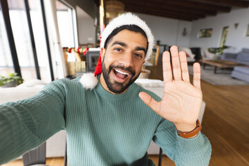 Happy biracial man with santa hat doing selfie, waving hand and smiling at christmas at home