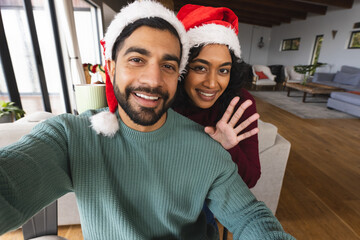 Happy biracial couple with santa hats doing selfie, waving hands and laughing at christmas at home