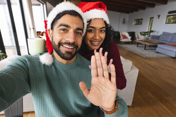 Happy biracial couple with santa hats doing selfie, waving hands and laughing at christmas at home